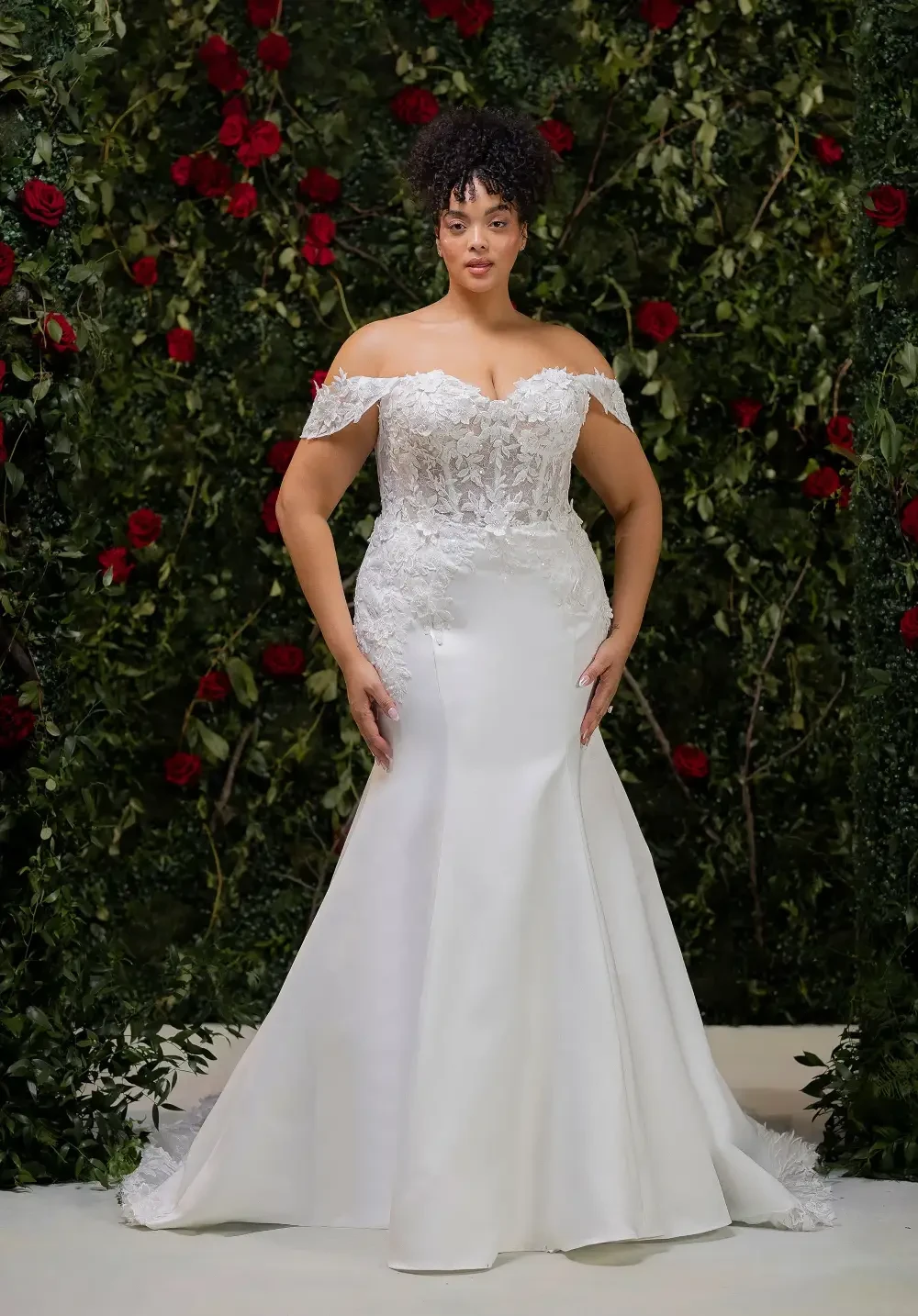 A woman wearing an elegant off-shoulder bridal gown with intricate lace detailing and a fitted silhouette, standing in front of a backdrop of lush greenery and red roses.