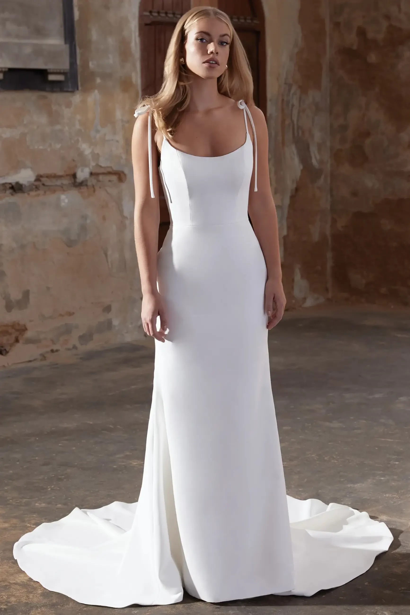 Model is wearing a simple and elegant scoop-neck wedding gown with a v-back and delicate bows on each shoulder.