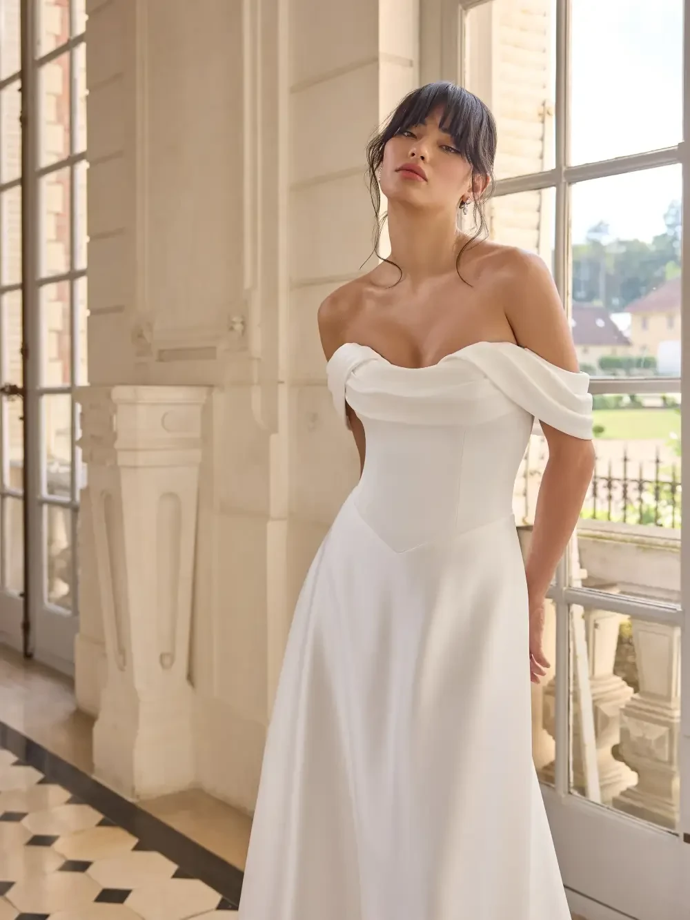 A model wearing a white off-shoulder gown standing in a well-lit, elegant indoor space with large windows.