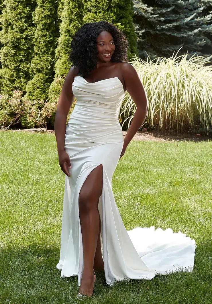 Model is wearing a plus size strapless fitted wedding dress with a thigh high slit and ruching on the bodice