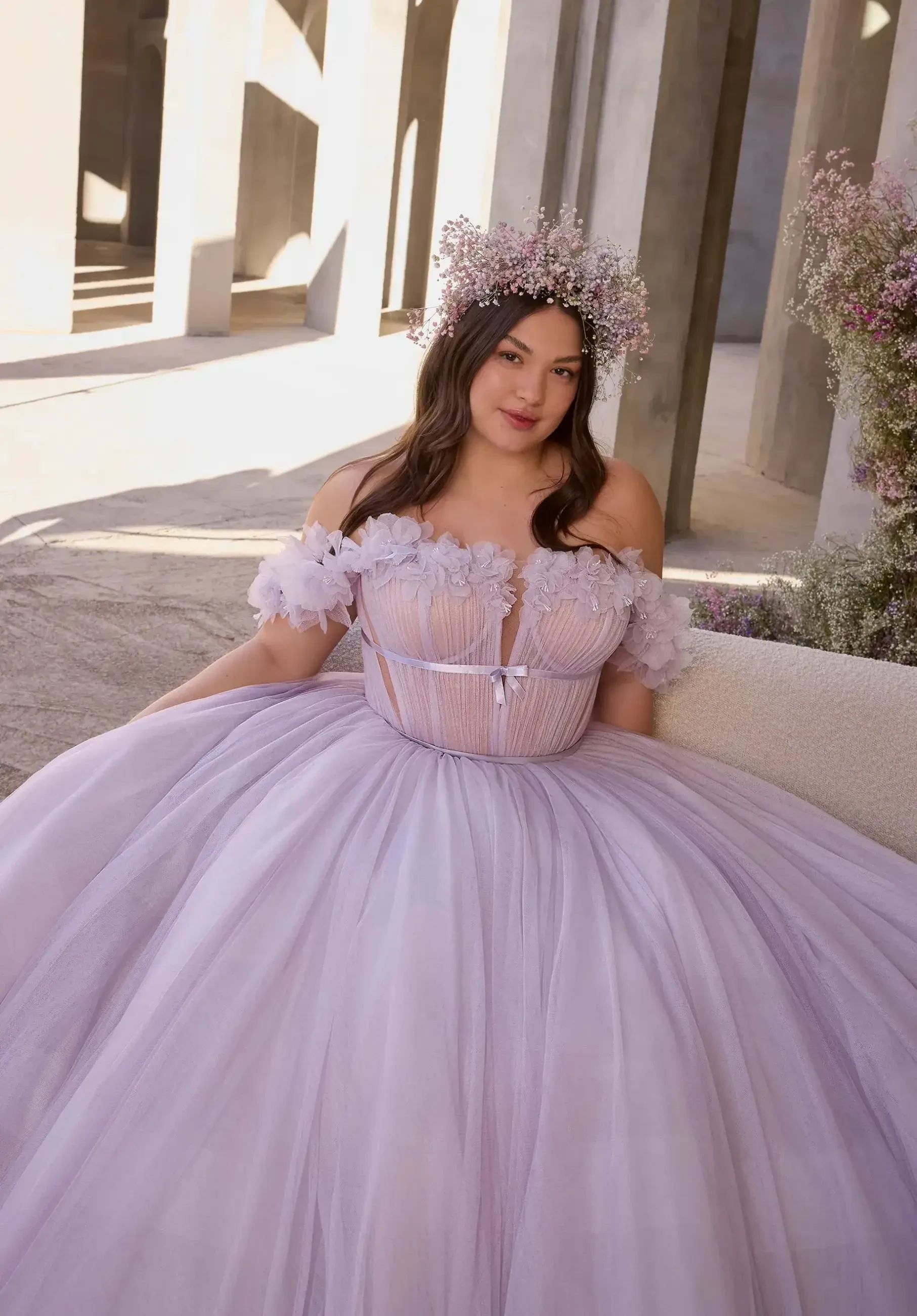 Young woman in a flowing lavender gown and floral crown sits in a sunlit, elegant setting with tall concrete columns, exuding a serene and joyful vibe.