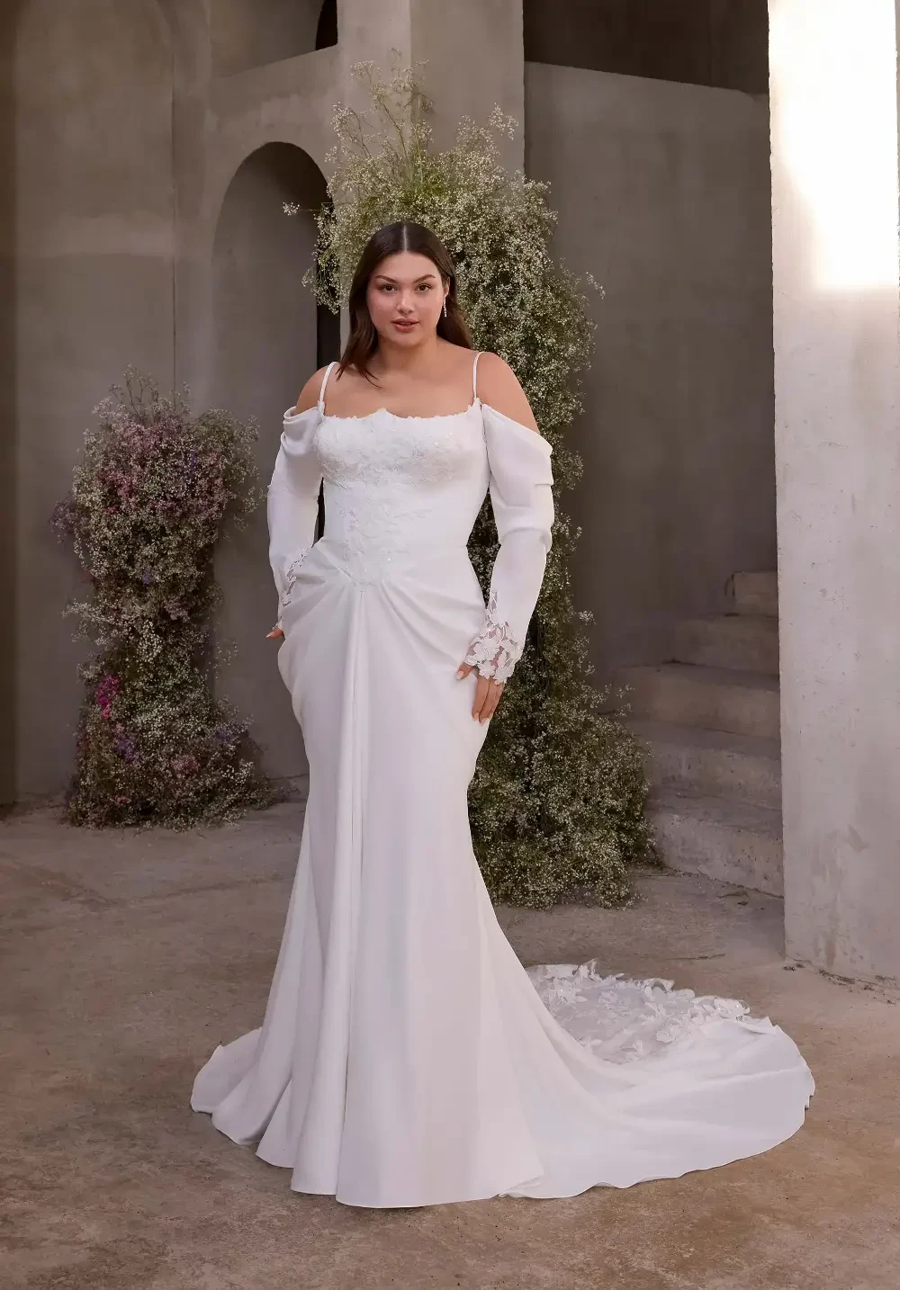 A woman stands in a white wedding dress with off-the-shoulder sleeves, surrounded by a rustic, outdoor setting featuring stone architecture and greenery.