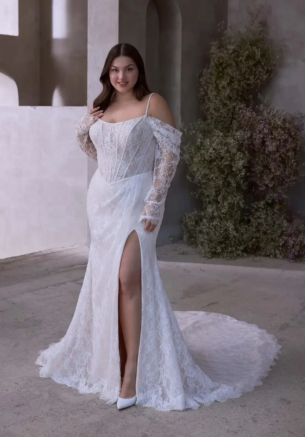 A model wearing a lace wedding gown with an off-the-shoulder design and a high slit, standing in a softly lit, minimalist setting.