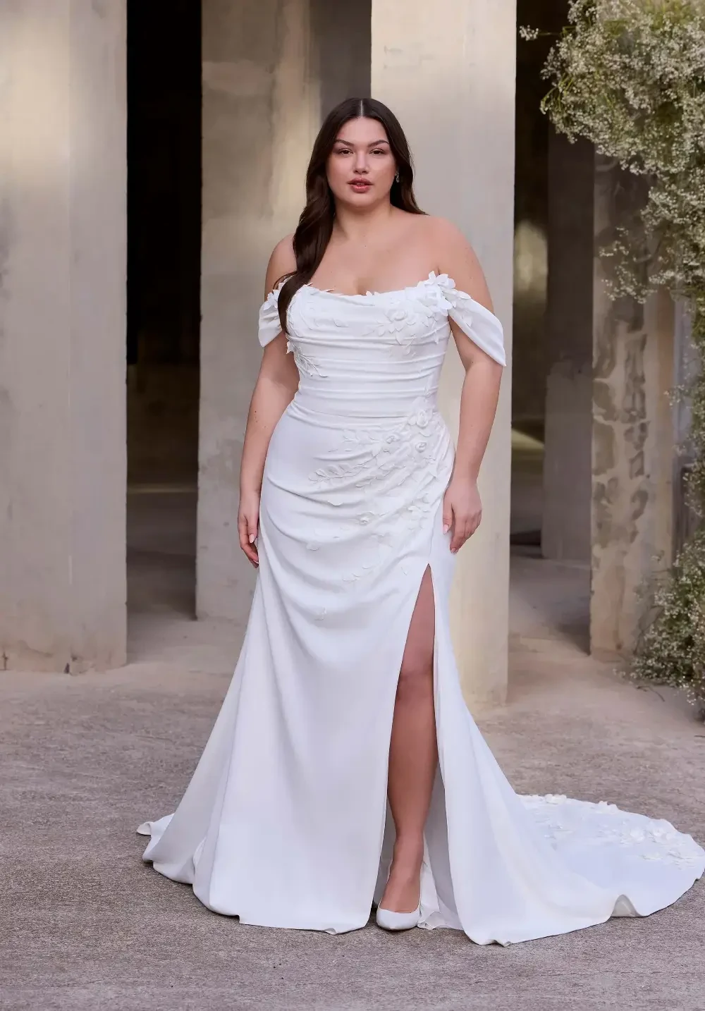 A woman wearing a white off-the-shoulder wedding dress with a thigh-high slit, standing in a minimalist outdoor setting with concrete pillars.