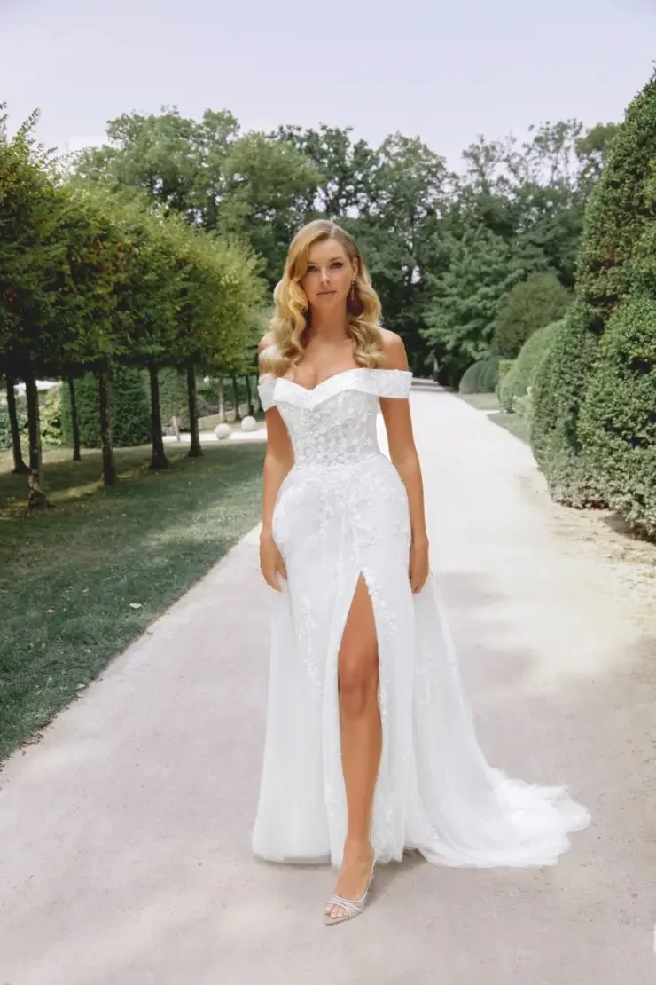 A woman in a strapless white wedding dress with a thigh-high slit stands on a garden path lined with manicured trees, exuding elegance and confidence.