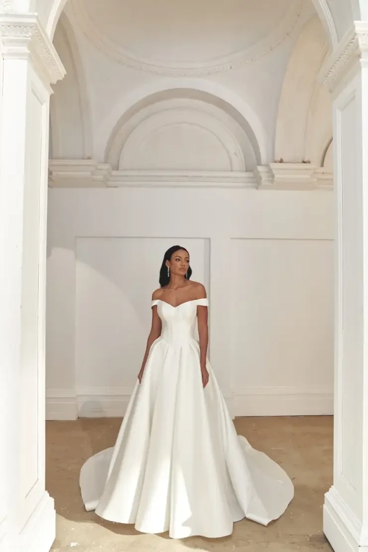 A woman in an elegant, off-the-shoulder white wedding dress stands in a grand, arched hallway. The setting exudes elegance and tranquility.
