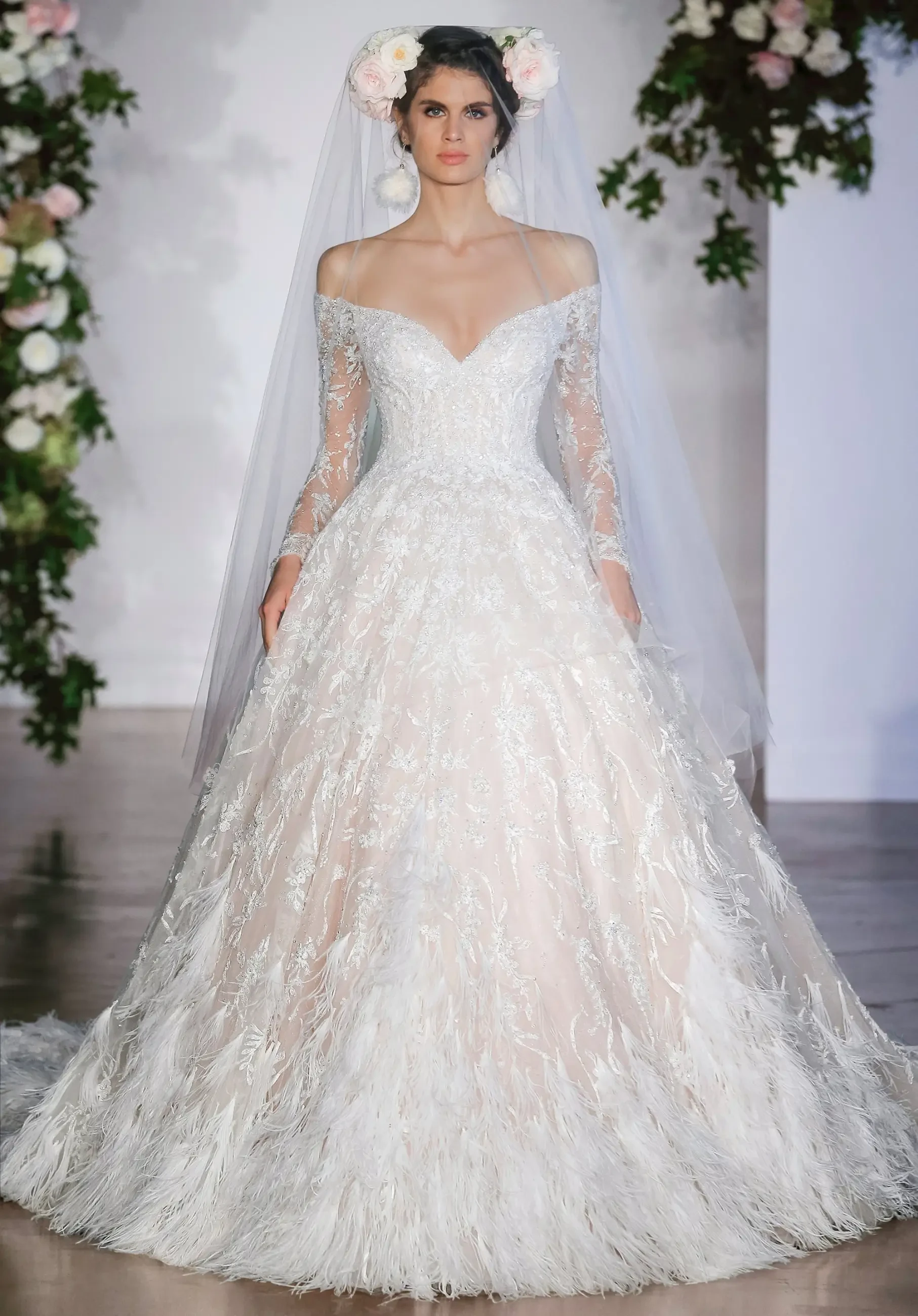 A model wearing an elegant off-the-shoulder wedding dress with long sheer sleeves, embellished with intricate lace and feather-like details, walking down a runway adorned with floral decorations.