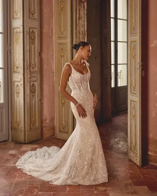 Bride in a shimmering white gown stands gracefully in an ornate room with tall, decorative doors, exuding elegance and serenity.