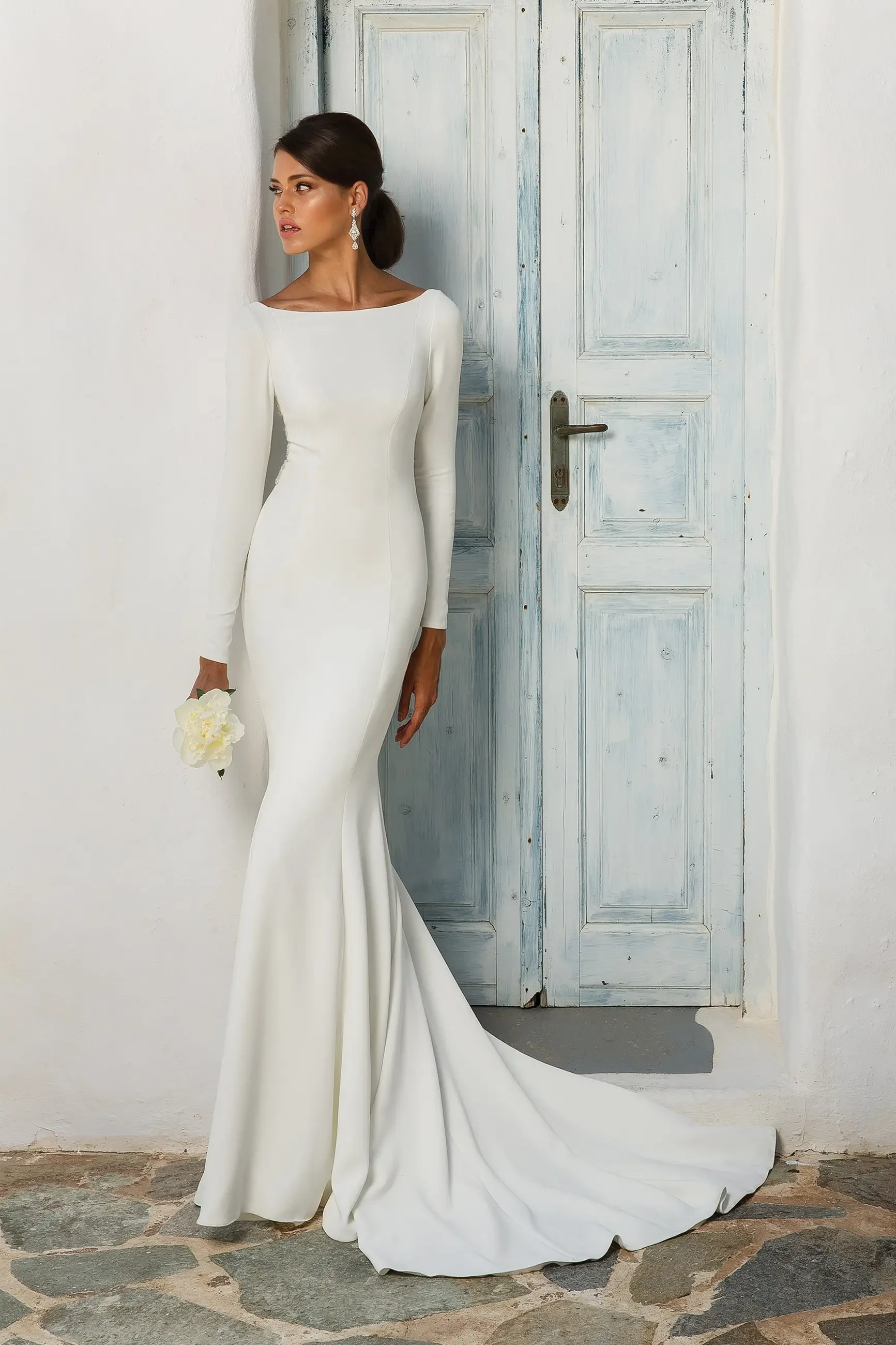 A woman in a long-sleeve white wedding dress stands gracefully next to a wooden door, holding a small bouquet.