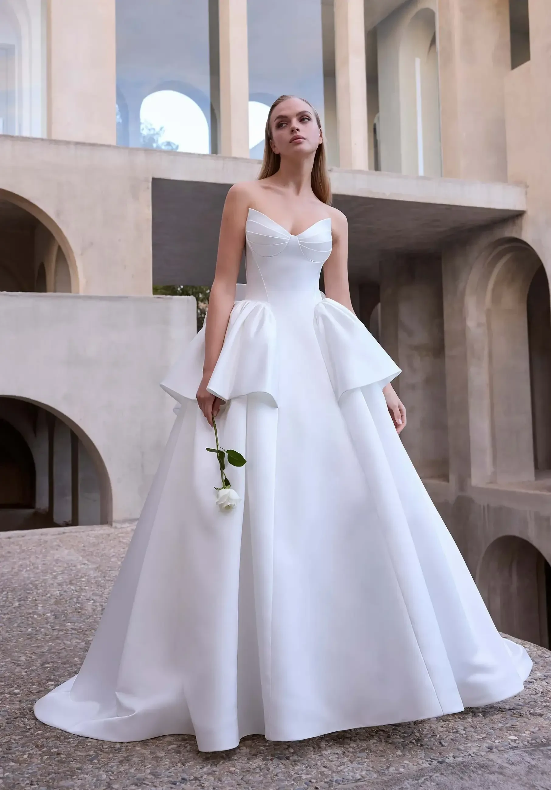 Model is wearing a satin A-line wedding dress with a cat-eye bodice, cascading ruffle peplum skirt, and a back bow.