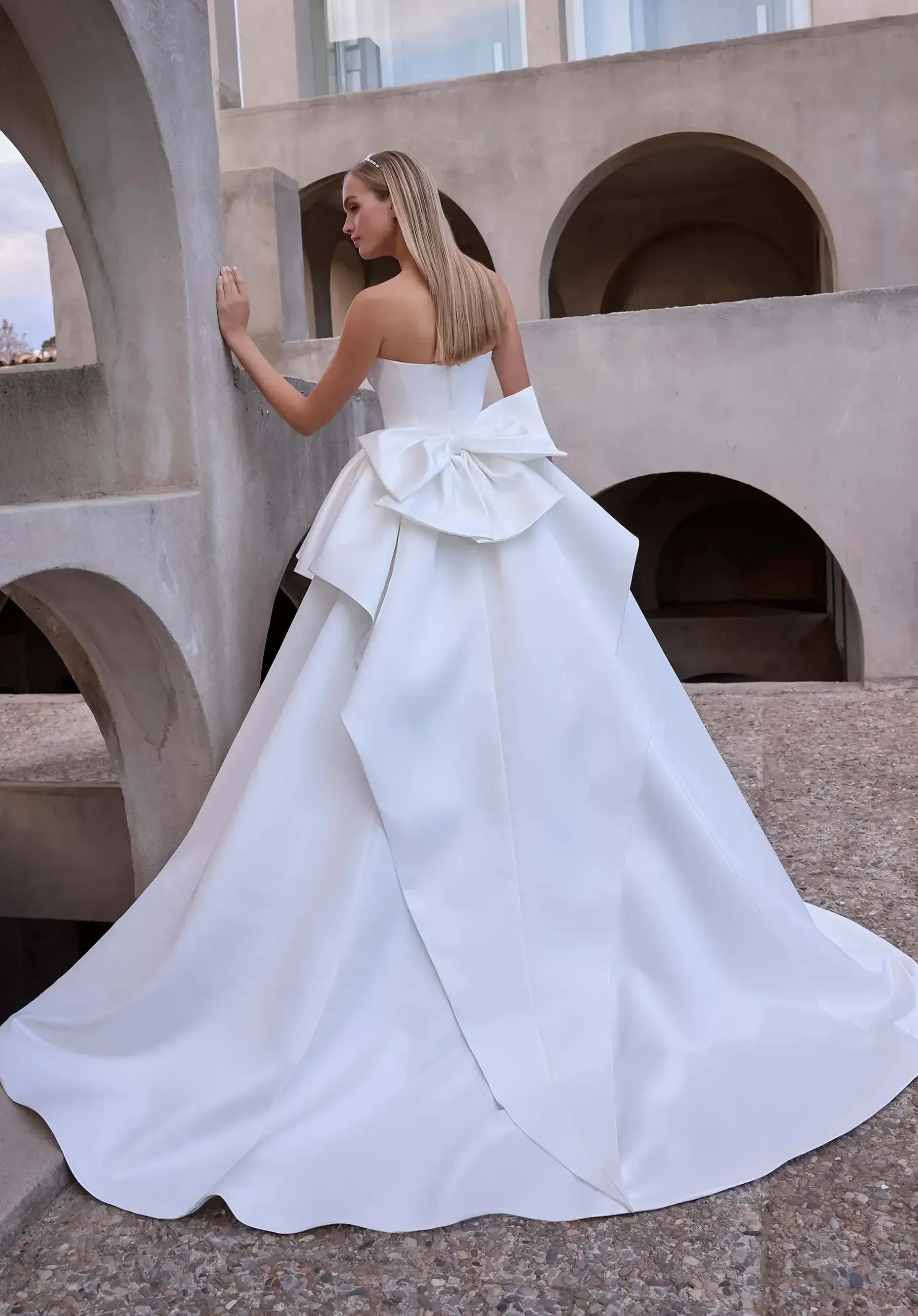 Model is wearing a satin A-line wedding dress with a cat-eye bodice, cascading ruffle peplum skirt, and a back bow.