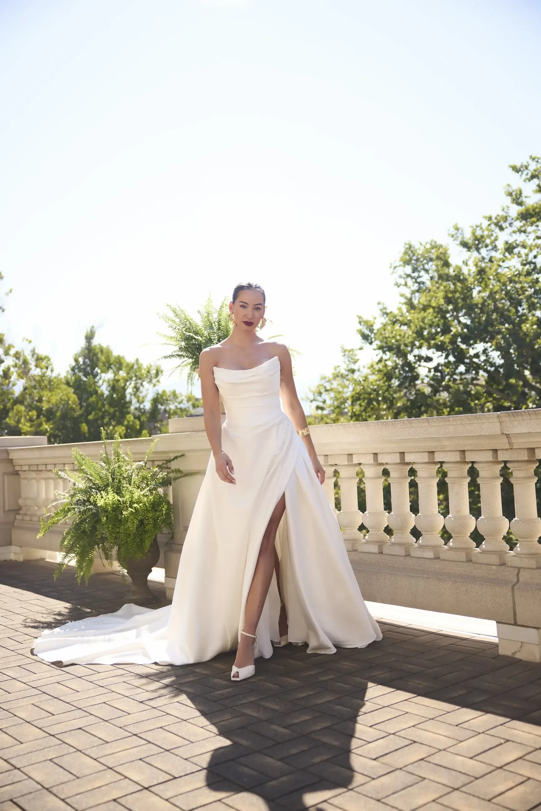 Bride in an elegant, off-shoulder white gown with a thigh-high slit stands on a sunny terrace, flanked by green ferns and bright trees, exuding grace.