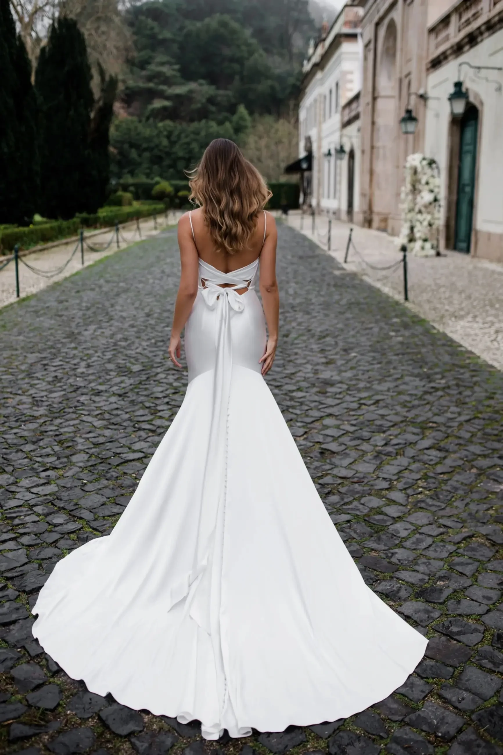 A woman in a flowing white wedding dress with a bow walks on a cobblestone path. Trees and a historic building create an elegant, serene backdrop.