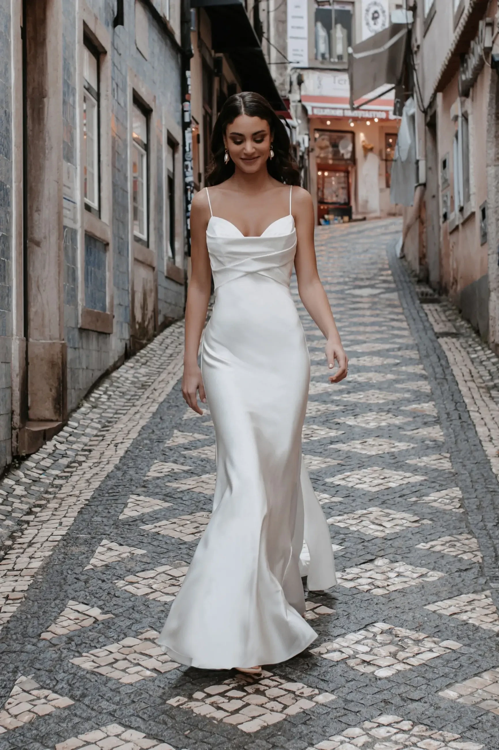 A woman in a flowing white dress walks gracefully down a narrow, cobblestone street lined with quaint, colorful buildings, exuding elegance and serenity.