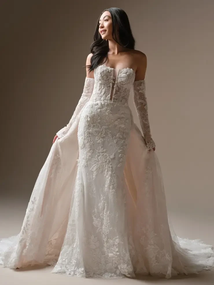 A model poses in an elegant, off-the-shoulder wedding gown with intricate lace detailing and long, sheer sleeves. The dress has a fitted bodice and a flowing skirt that creates a romantic silhouette.