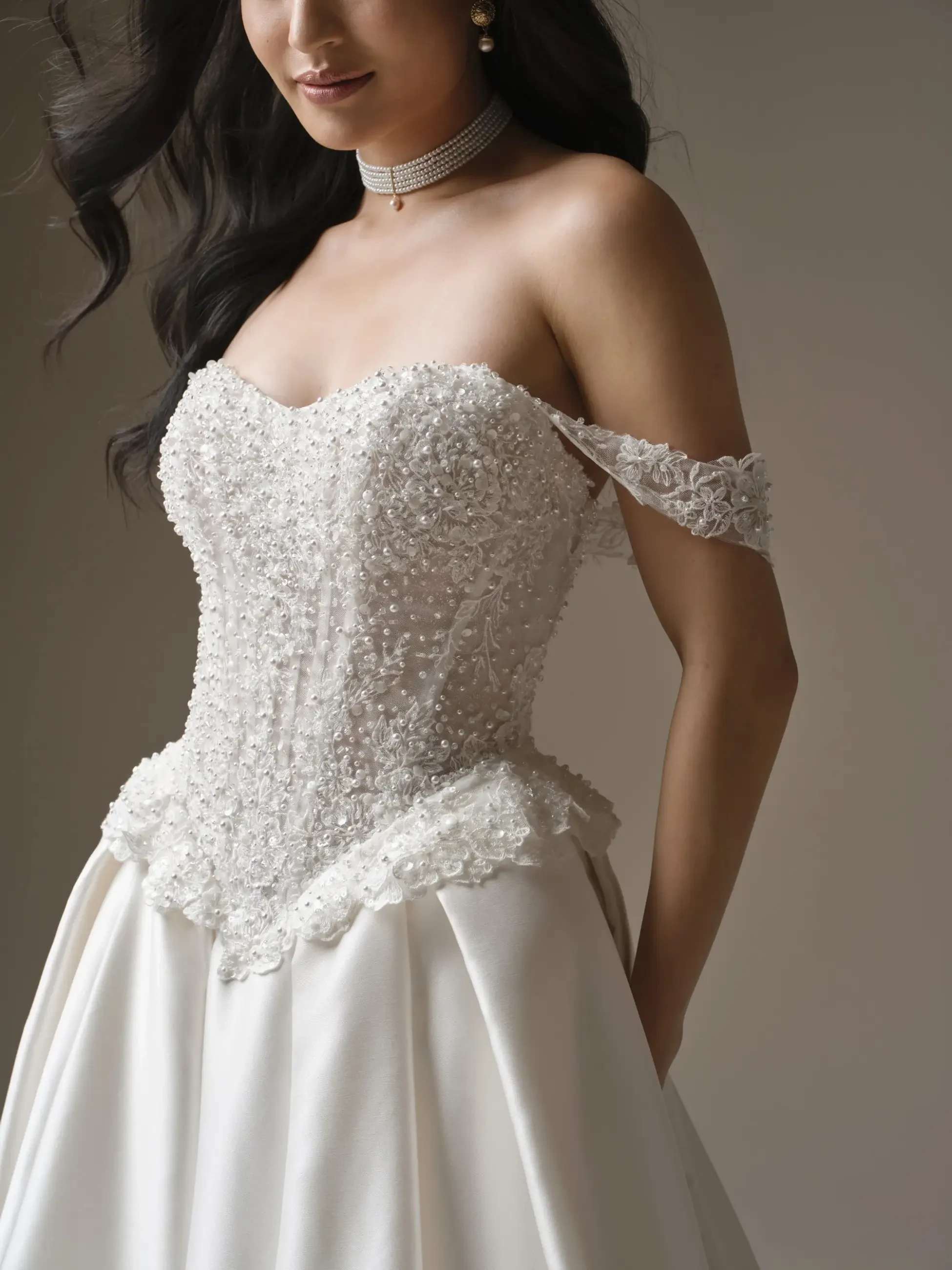 A bride in an off-shoulder wedding dress with intricate pearl embroidery, exudes elegance. Her wavy hair and subtle jewelry add a classic touch.