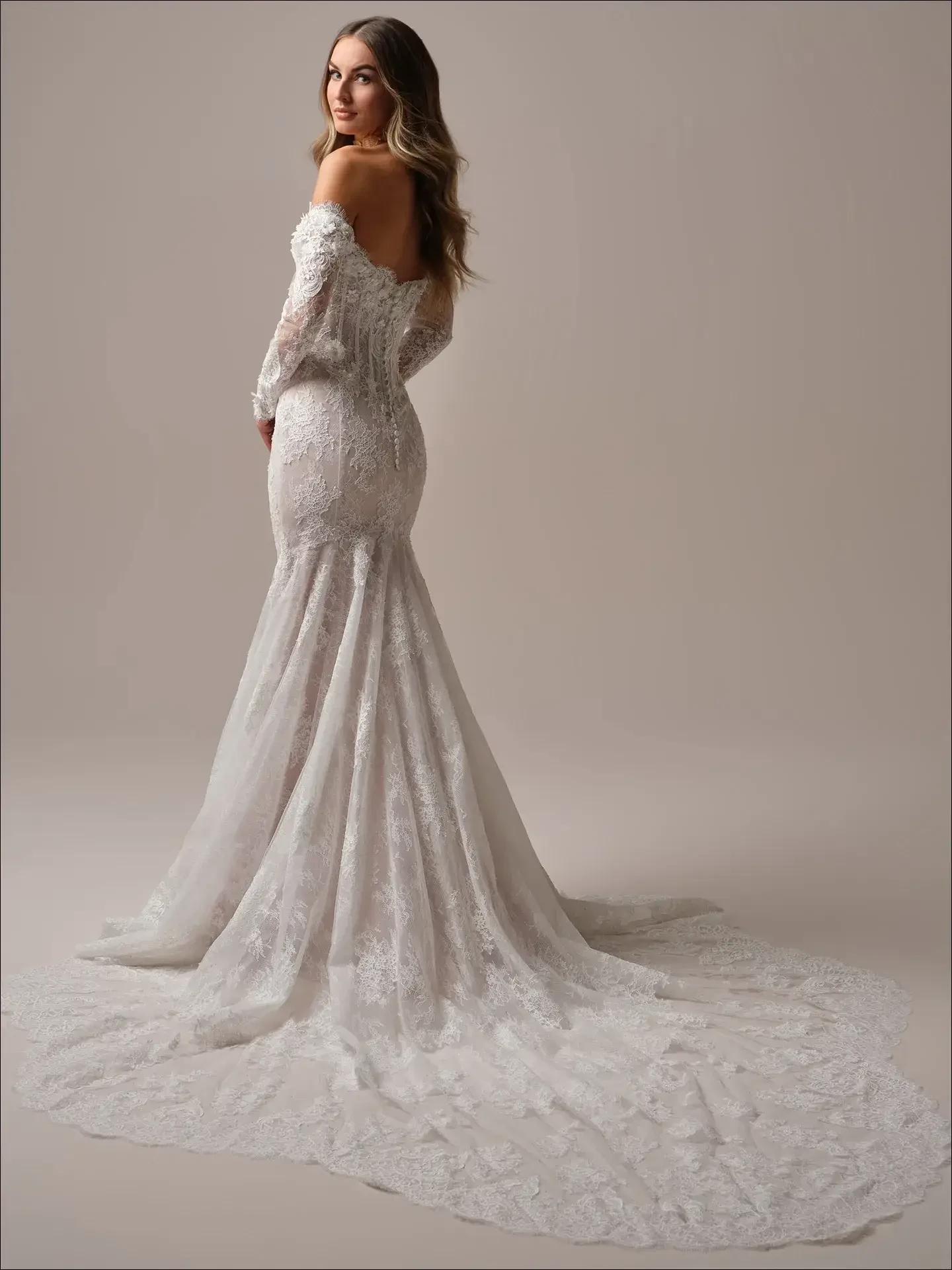A woman in an elegant off-the-shoulder, lace mermaid wedding dress with long sleeves and a long train, set against a neutral background.