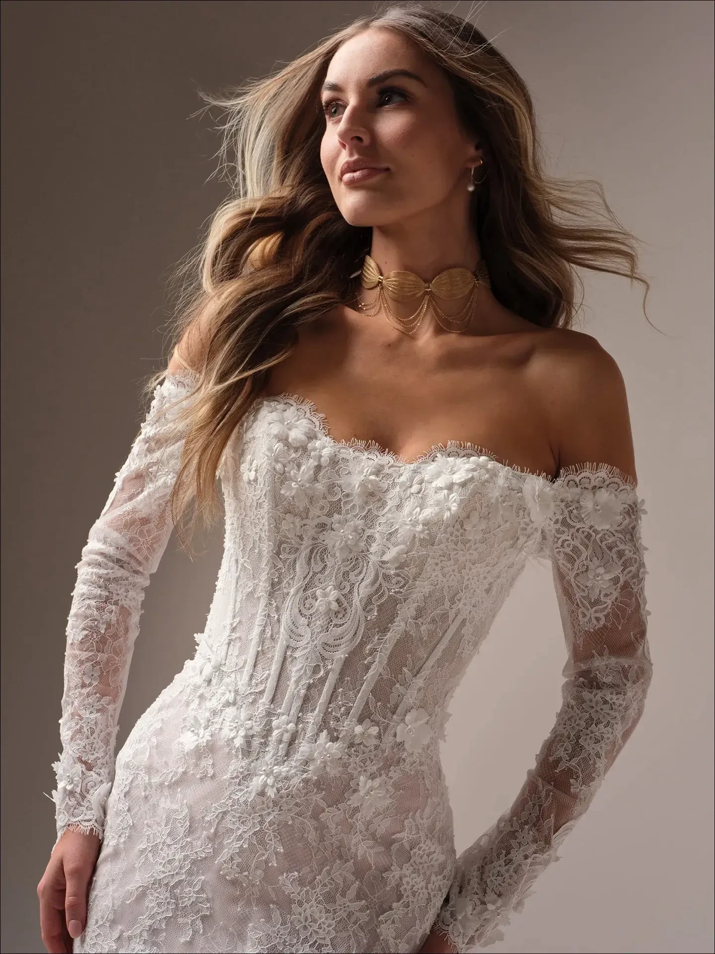 A woman in a lace off-shoulder wedding dress with intricate floral patterns. She looks confidently upward, her hair gently flowing, wearing a gold choker.