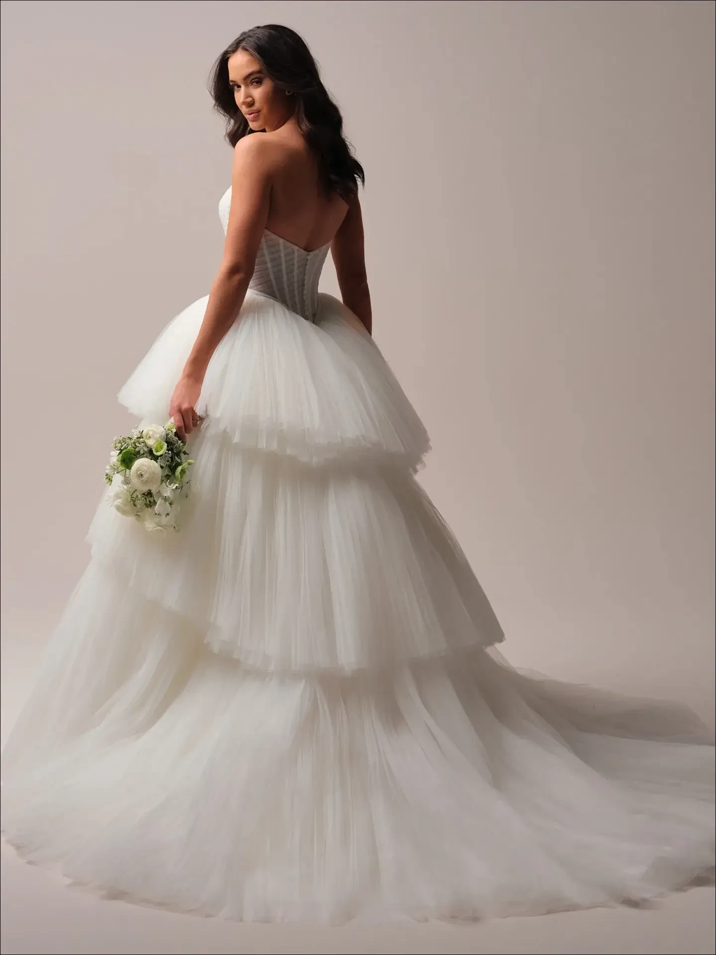 A bride in a tiered, strapless wedding gown holds a bouquet of white and green flowers. She stands gracefully, exuding elegance and serenity.