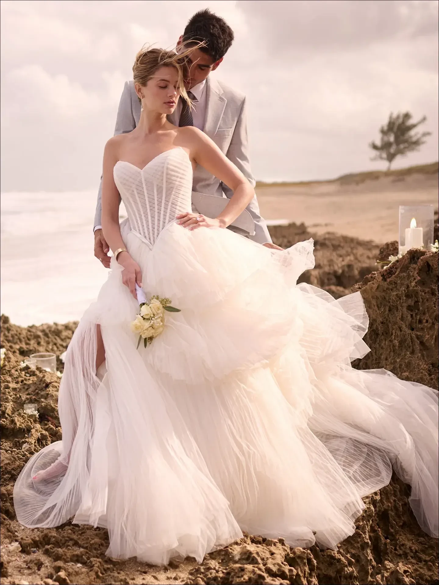 Bride in a strapless, flowing wedding gown stands on rocky beach, groom in light suit gently embraces her. Serene and romantic atmosphere.